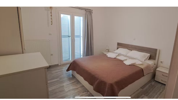 A bedroom with a bed, bedside tables, a wardrobe, and a window with curtains. - Central Paroikia Apartment - 10min walk to Port and Beach! (Paros)