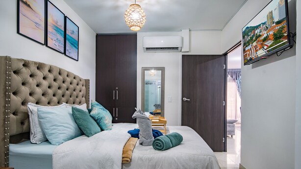 Ein Schlafzimmer mit einer gesteppten Kopfteil, einem Bett mit weißer Bettwäsche, mehreren Kissen, einem aufgerollten Handtuch, einem Holznachttisch, einem Fernseher und einer Wand-Klimaanlage.