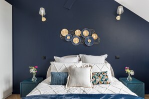 Une chambre à coucher avec un lit, des œuvres d’art décoratives au mur, deux tables de chevet avec des vases et un mur bleu foncé.