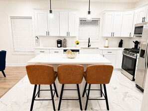A modern kitchen with white cabinets, a central island with bar stools, a stove, and a refrigerator. - Fire Pit Home by Mercedes-Benz Stadium (Atlanta)