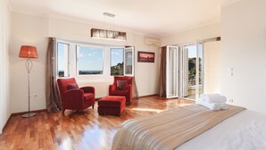 A bedroom with a large bed, a red armchair, a small ottoman, a floor lamp, and a view of the outdoors through a sliding door. - Villa Amarandes With Private Pool (Rhodes)