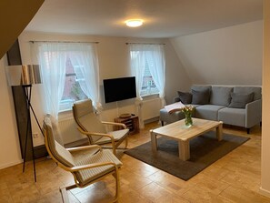 Ein Wohnzimmer mit einer Couch, zwei Sesseln, einem Couchtisch, einem Fernseher und einer Heizung.