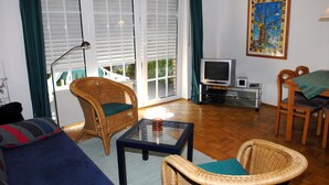 Una sala de estar con un sofá, dos sillas de mimbre, una mesa de centro de vidrio, un televisor y un cuadro en la pared.