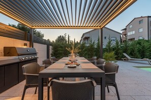 Un patio exterior con una parrilla, mesa de comedor y sillas, rodeado de follaje y edificios.