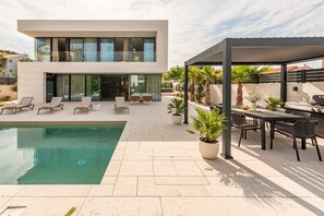 Una casa moderna de dos pisos con piscina, mobiliario exterior y un patio cubierto.