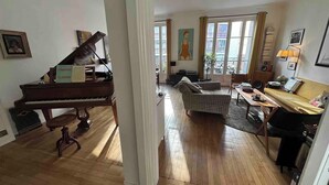 En stue med et flygell, en sofa, et spisebord og udsigt til en balkon gennem en døråbning.