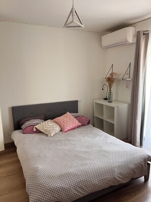 A bedroom with a bed, a bedside table with a vase, and an air conditioning unit. - Between lake and sea (Clermont-l'Hérault)