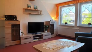 Ein Wohnzimmer mit Flachbildfernseher, Holzausbau, einem Esstisch mit dekorativer Tischdecke und einem Fenster mit Vorhängen.