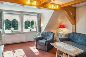 Una habitación acogedora con dos sillones, una mesa de madera con camino de encaje, una lámpara roja y una ventana con cortinas.