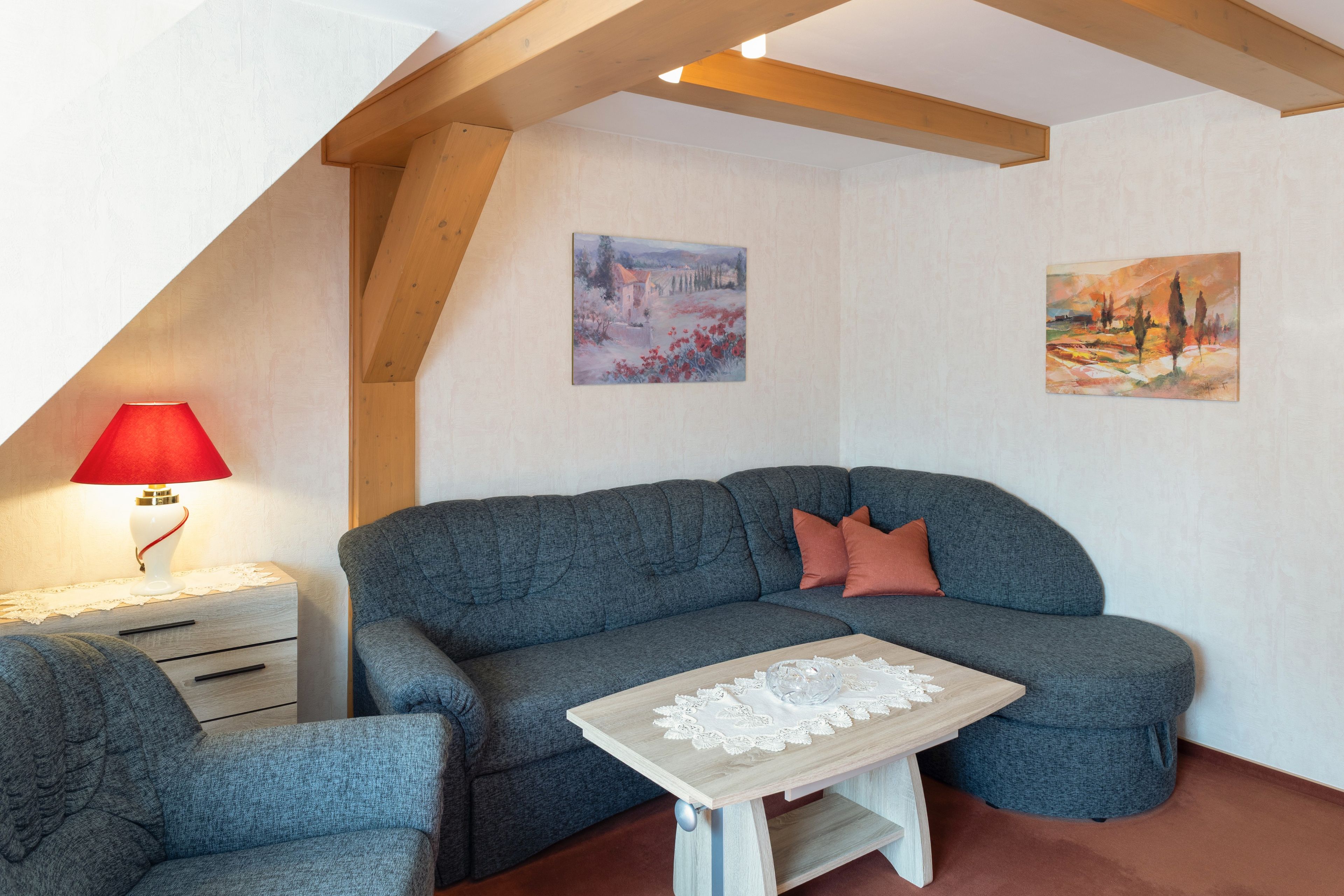 A cozy living room with a grey sofa, a small table with a lace tablecloth, and two paintings on the wall.