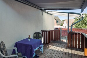 Un balcón techado al aire libre con una mesa y sillas, una planta en maceta y vistas del barrio.
