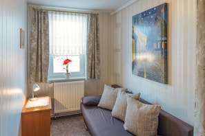 A room with a sofa, a table with a lamp, a window with curtains, a radiator, and a painting on the wall.