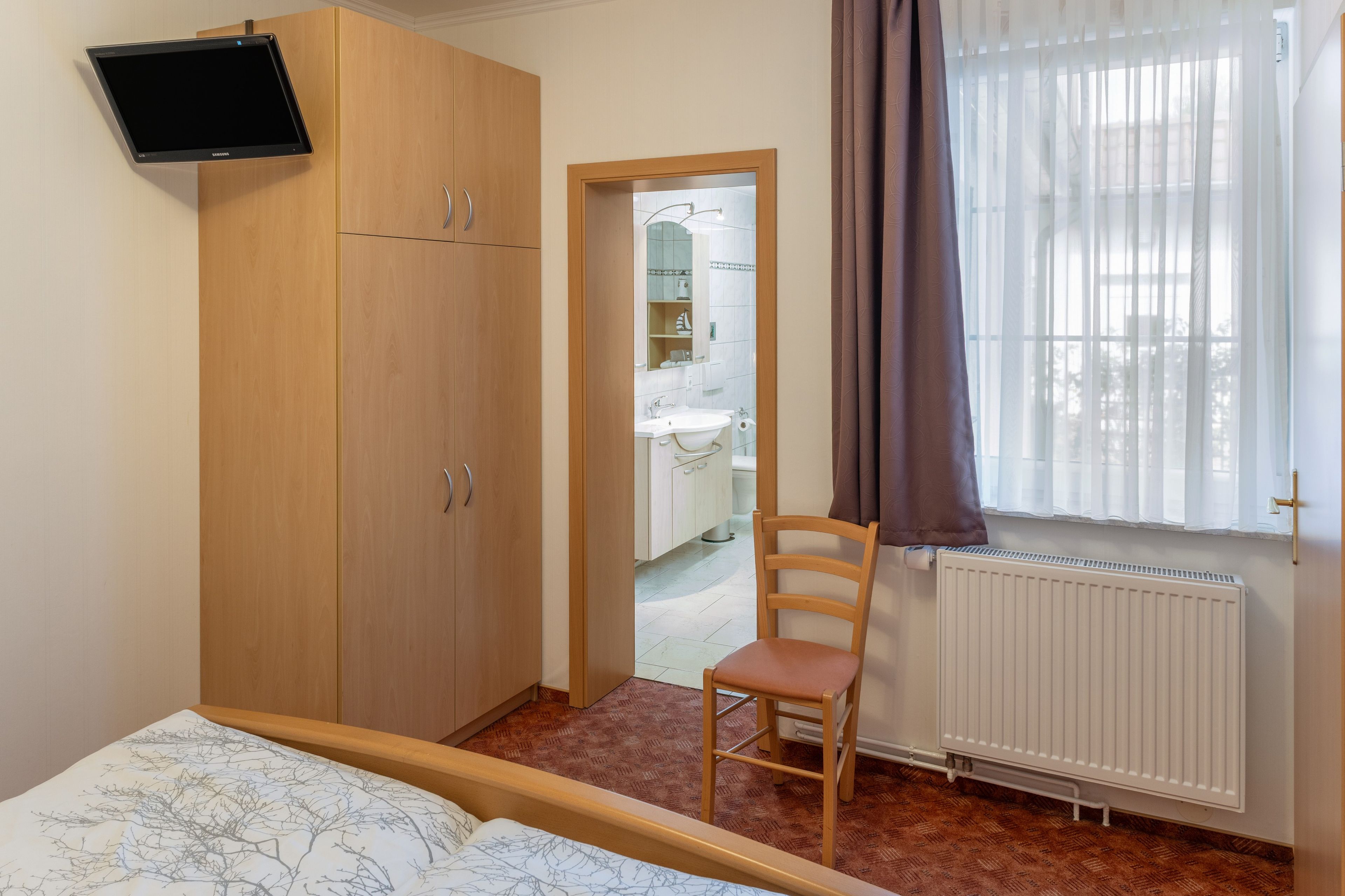 A bedroom with a bed, a wooden wardrobe, a chair, a television mounted on the wall, and a window with curtains.