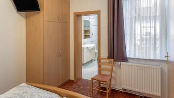 Ein Schlafzimmer mit einem Bett, einem Holzschrank, einem Sessel, einem an der Wand befestigten Fernseher und einem Fenster mit Vorhängen.