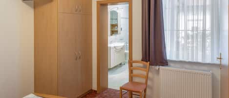 A bedroom with a bed, a wooden wardrobe, a chair, a television mounted on the wall, and a window with curtains.