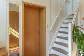 Una puerta de madera con manija de latón, una escalera con barandilla blanca y una planta en maceta a la derecha.