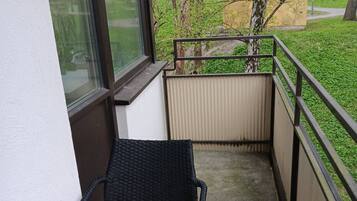 En balkong med en svart flätstol, utsikt över en grön park och en byggnad i bakgrunden.