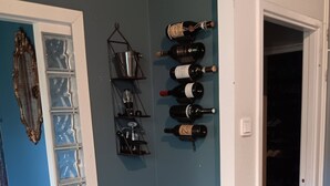 A room with a wooden stool, a metal shelf with books, a wine rack, and a door leading to another room.