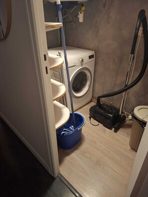 A compact laundry room with a washing machine, a vacuum cleaner, and a blue bucket. - Paradis d'Azur: 1-bedroom apt facing the sea, a/c (Menton)