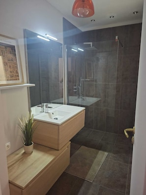 A modern bathroom with a large mirror, a white sink, a shower, and a potted plant.