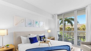 Ein Hotelzimmer mit einem Bett, Kunstwerken an der Wand, einem Balkon mit Stühlen und Blick auf Palmen.