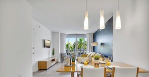 Una sala de estar moderna con comedor, ventanas amplias, un televisor de pantalla plana y vistas a palmeras.