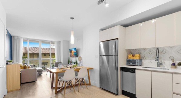 A modern kitchen with stainless steel appliances, a dining area with a wooden table and chairs, and a large window offering a view of the outside. - Beachwalk Resort · 2BR · Intracoastal View (Hallandale Beach)