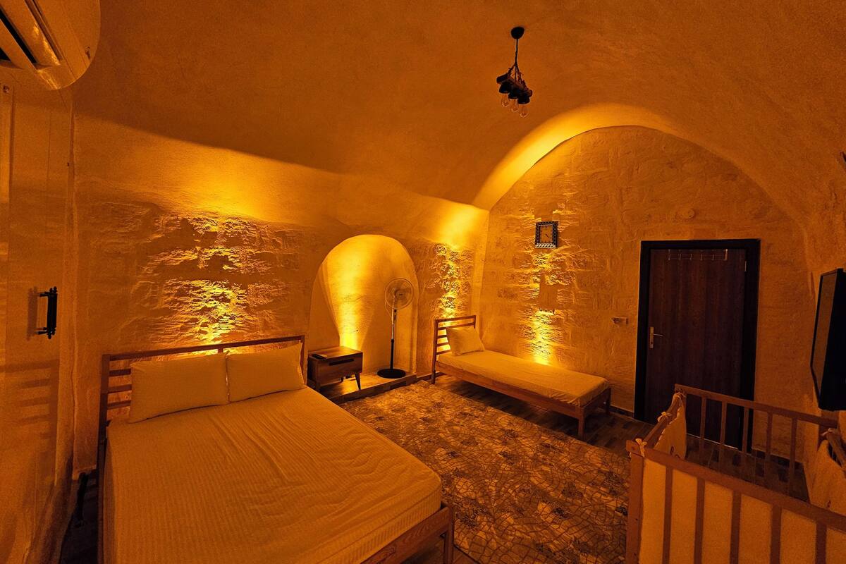 A cozy room with a bed, a bench, and a small table. There are two arched alcoves with lighting, a wooden door, and a ceiling fan.