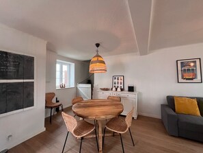 Una sala moderna con una mesa de comedor redonda de madera, sillas de mimbre, un sofá gris con un cojín color mostaza, un armario blanco y una lámpara colgante.