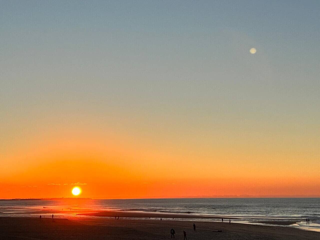 Escena de playa con el sol poniéndose sobre el océano, siluetas de personas en la arena y una luna visible en el cielo.