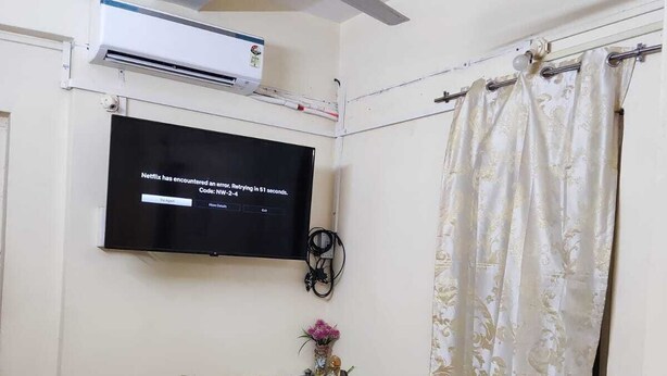 Una habitación con una televisión de pantalla plana mostrando un mensaje de error, un ventilador de techo, un equipo de aire acondicionado, una mesita con un jarrón y un suelo a cuadros.