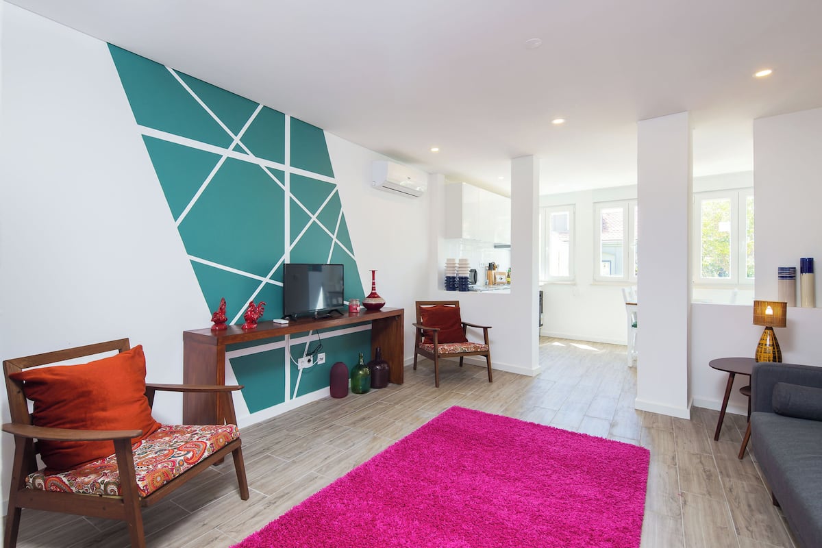 Un salon moderne avec un mur orné d’un motif géométrique turquoise, un téléviseur à écran plat, des meubles en bois, un tapis rose vif et une vue sur la cuisine.