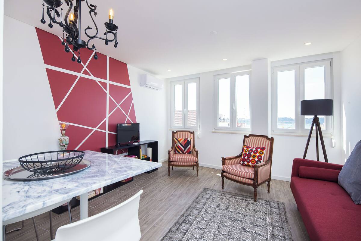 A modern living room with a dining table, chairs, a sofa, a chandelier, and large windows.