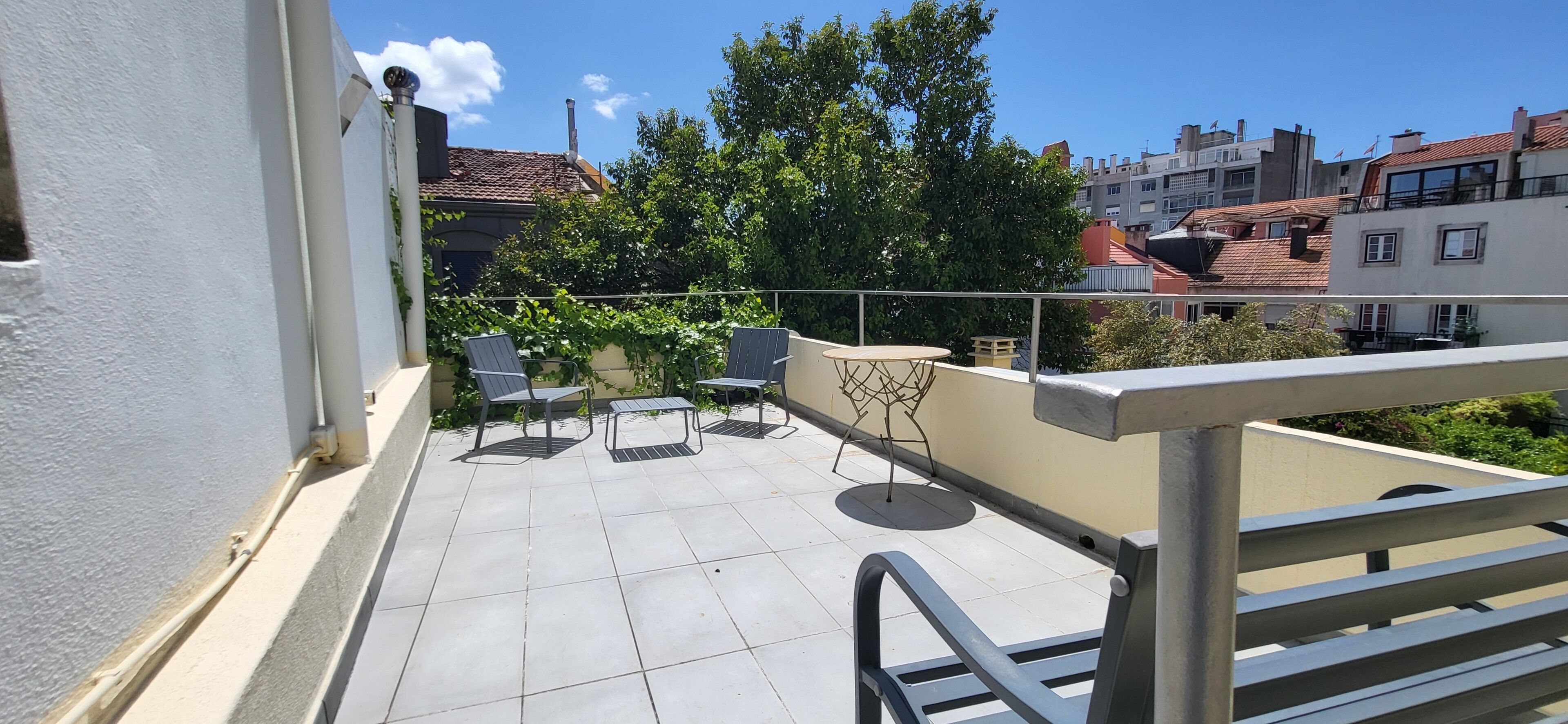 A balcony with chairs and a table, surrounded by buildings and trees.