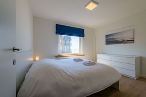 Ein Schlafzimmer mit einem Bett, einem Fenster mit Blick auf ein Gebäude, einem Bild an der Wand und einer weißen Kommode.