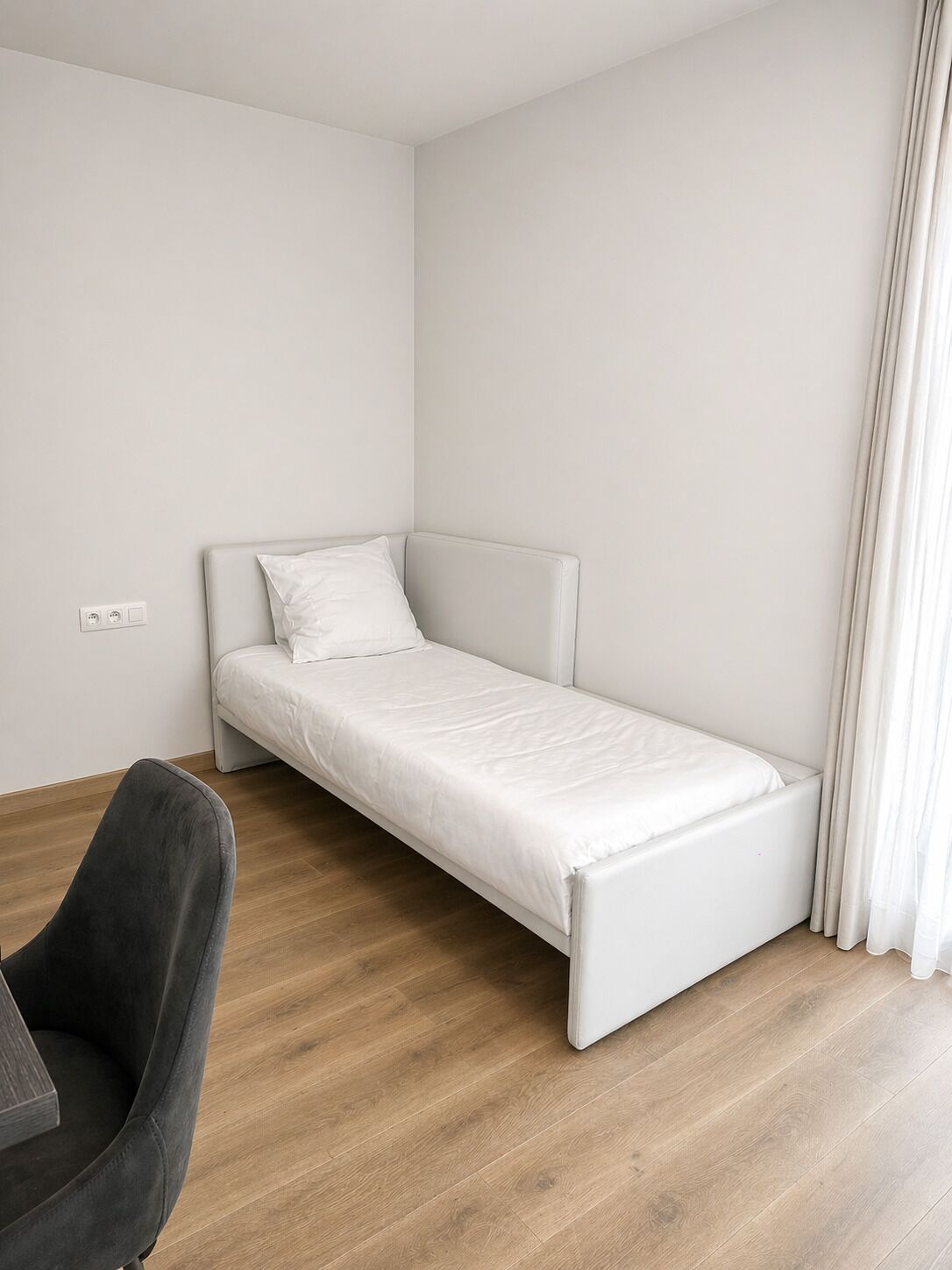 A small, single-bed room with a wooden floor, a grey chair, and a white bed.