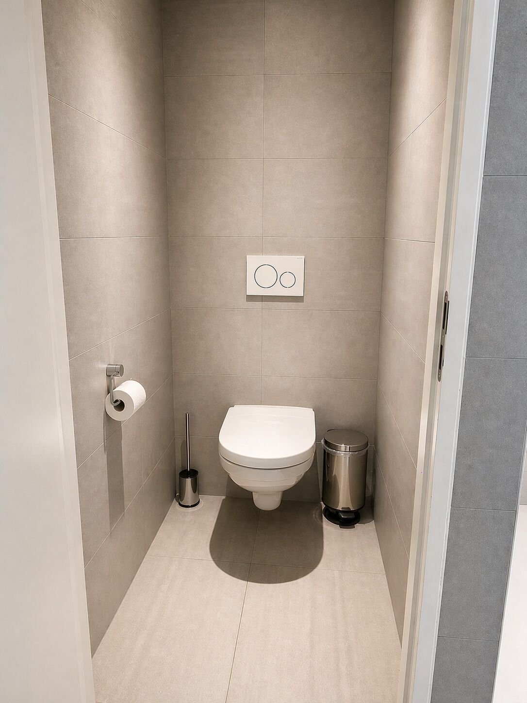 A bathroom with a toilet, a trash bin, a toilet paper holder, and a wall-mounted flush control panel.