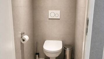 A bathroom with a toilet, a trash bin, a toilet paper holder, and a wall-mounted flush control panel.