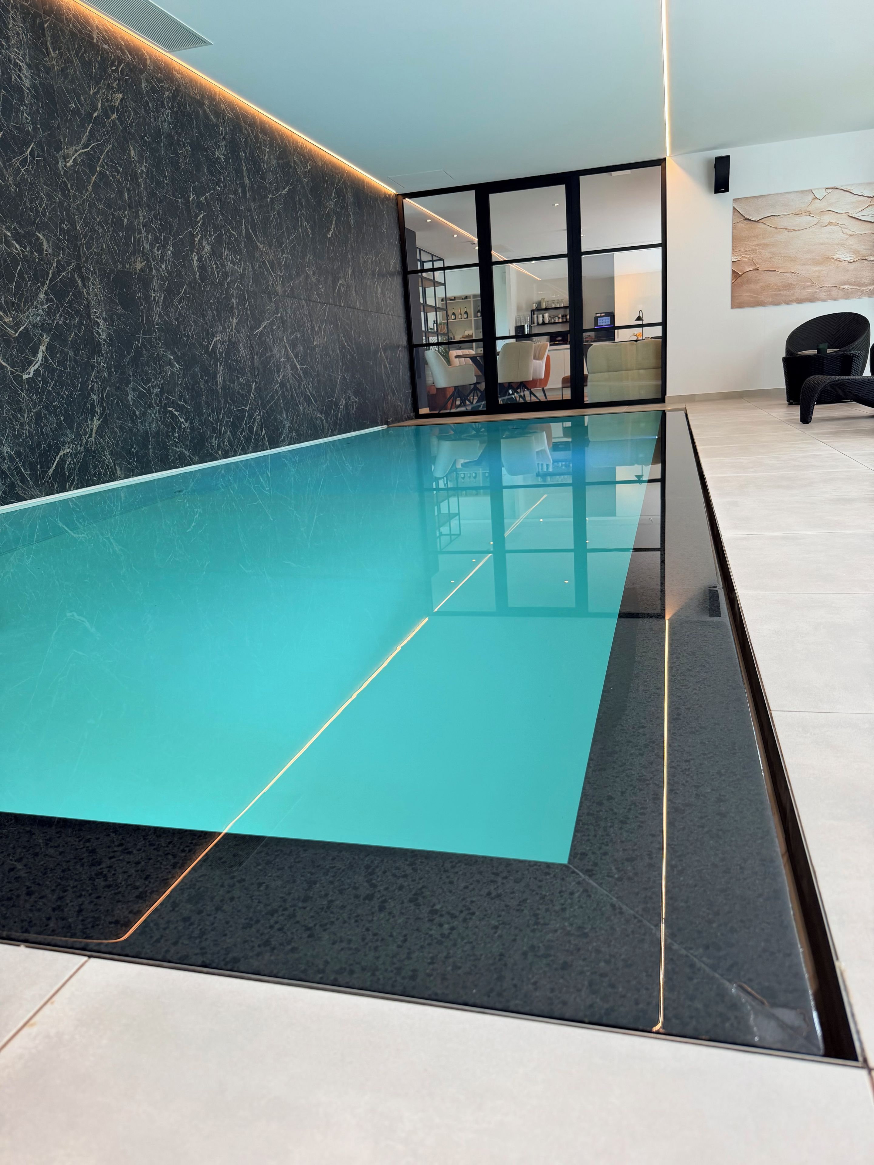 A modern indoor swimming pool with a glass bottom, surrounded by a dark marble wall and a tiled floor.