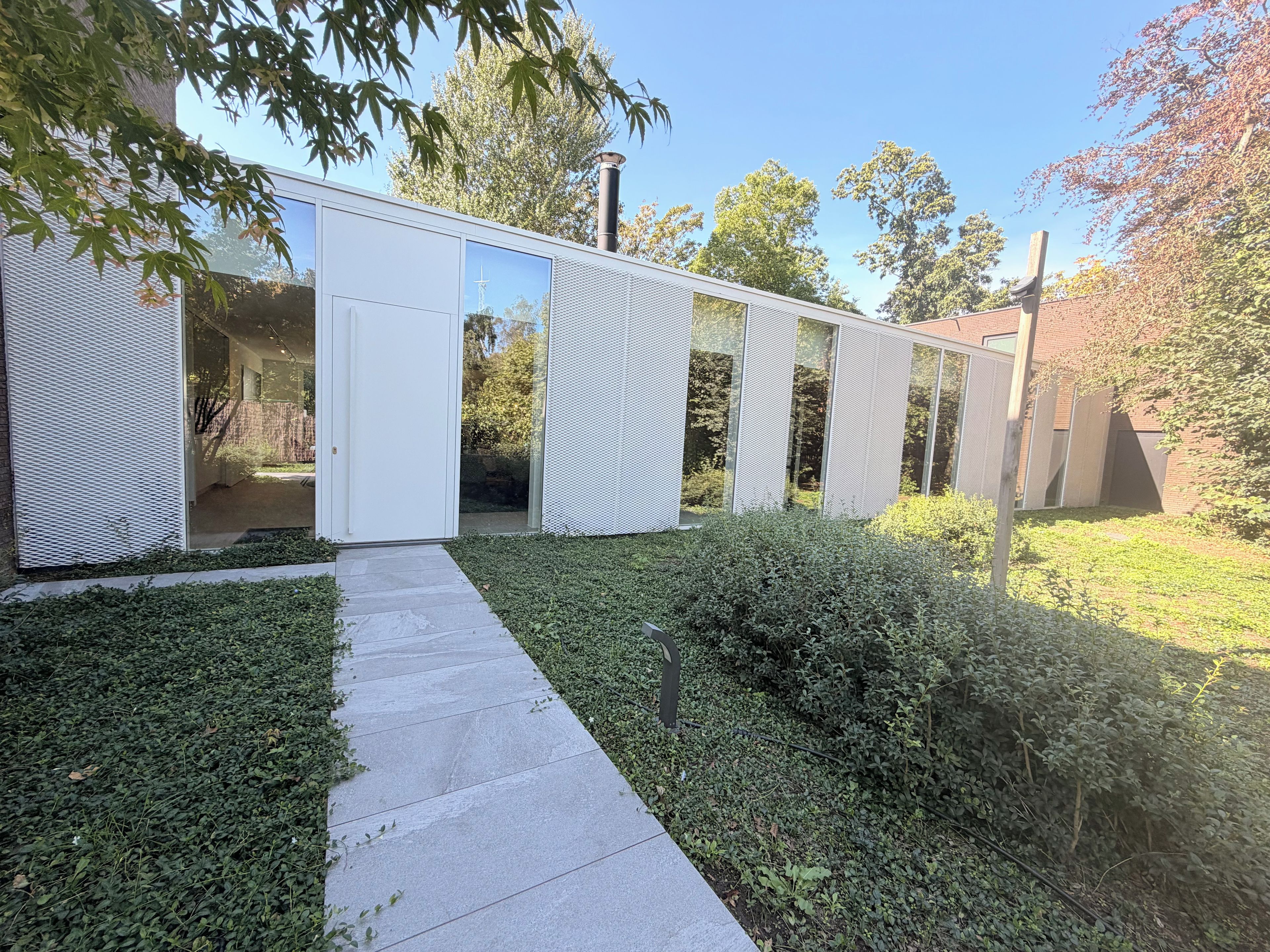 Un bâtiment moderne avec une entrée dotée d’une porte en verre, un chemin en béton et une végétation luxuriante tout autour.