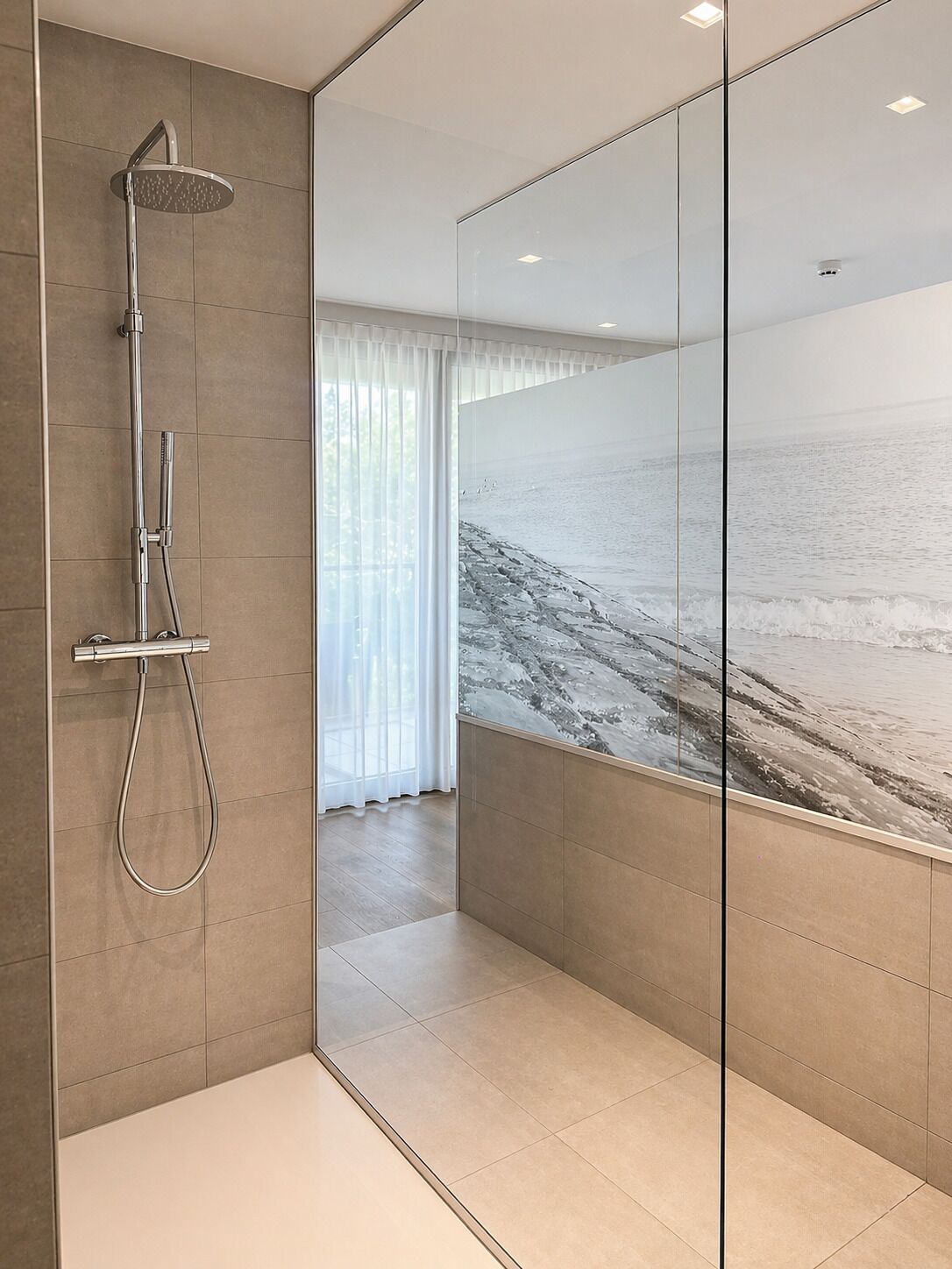 Une salle de bain moderne dotée d’une cabine de douche en verre, d’un pommeau de douche apparent et d’un grand mur en verre offrant une vue sur la plage.