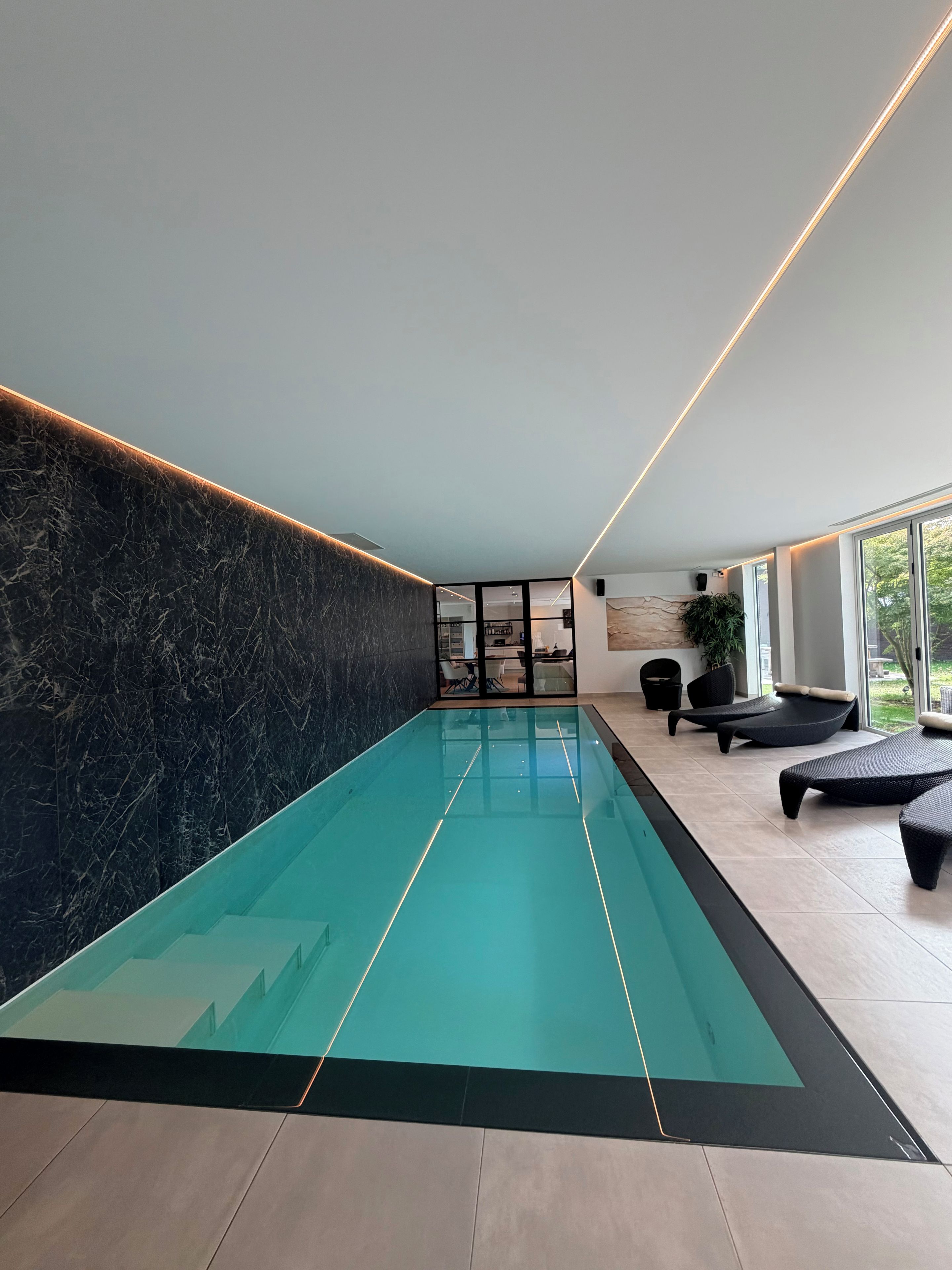 A modern indoor swimming pool with a glass bottom, surrounded by seating areas and a stone wall.