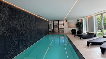 A modern indoor swimming pool with a glass bottom, surrounded by seating areas and a stone wall.