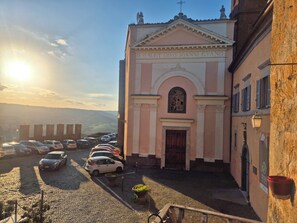 Una iglesia con fachada rosa, un patio empedrado, autos estacionados y un cielo despejado.