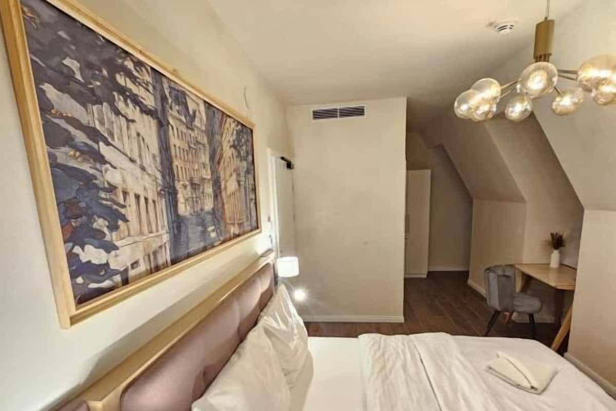 A hotel room with a bed, a nightstand, a wall-mounted lamp, a ceiling light fixture, and a large painting.