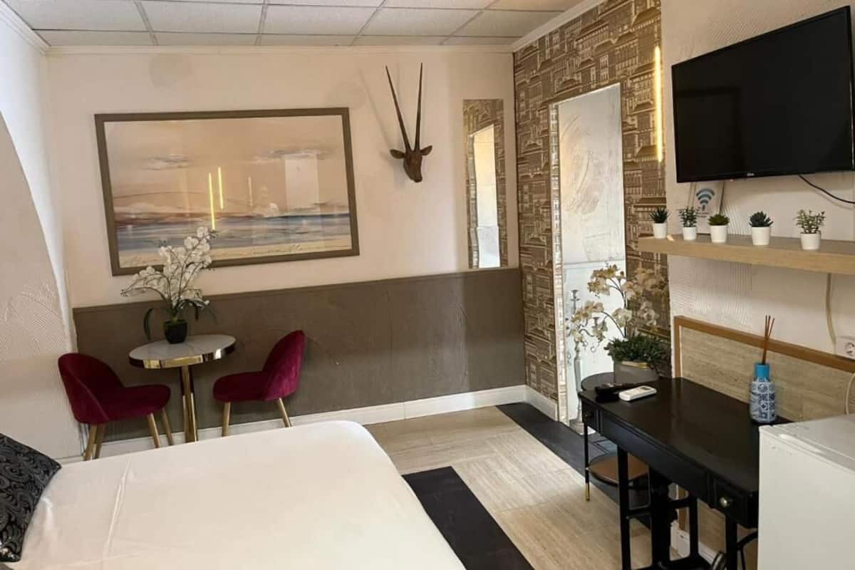 A hotel room with a bed, a desk, two chairs, a TV, a wall-mounted shelf with plants, and a decorative wall piece.