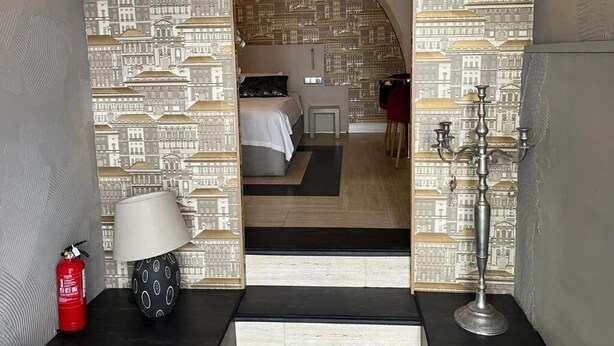 A hotel room with a patterned wallpaper, a bed, a bedside table with a lamp, a red fire extinguisher, and a metal stand with candles. - São Pedro Suítes (Faro)