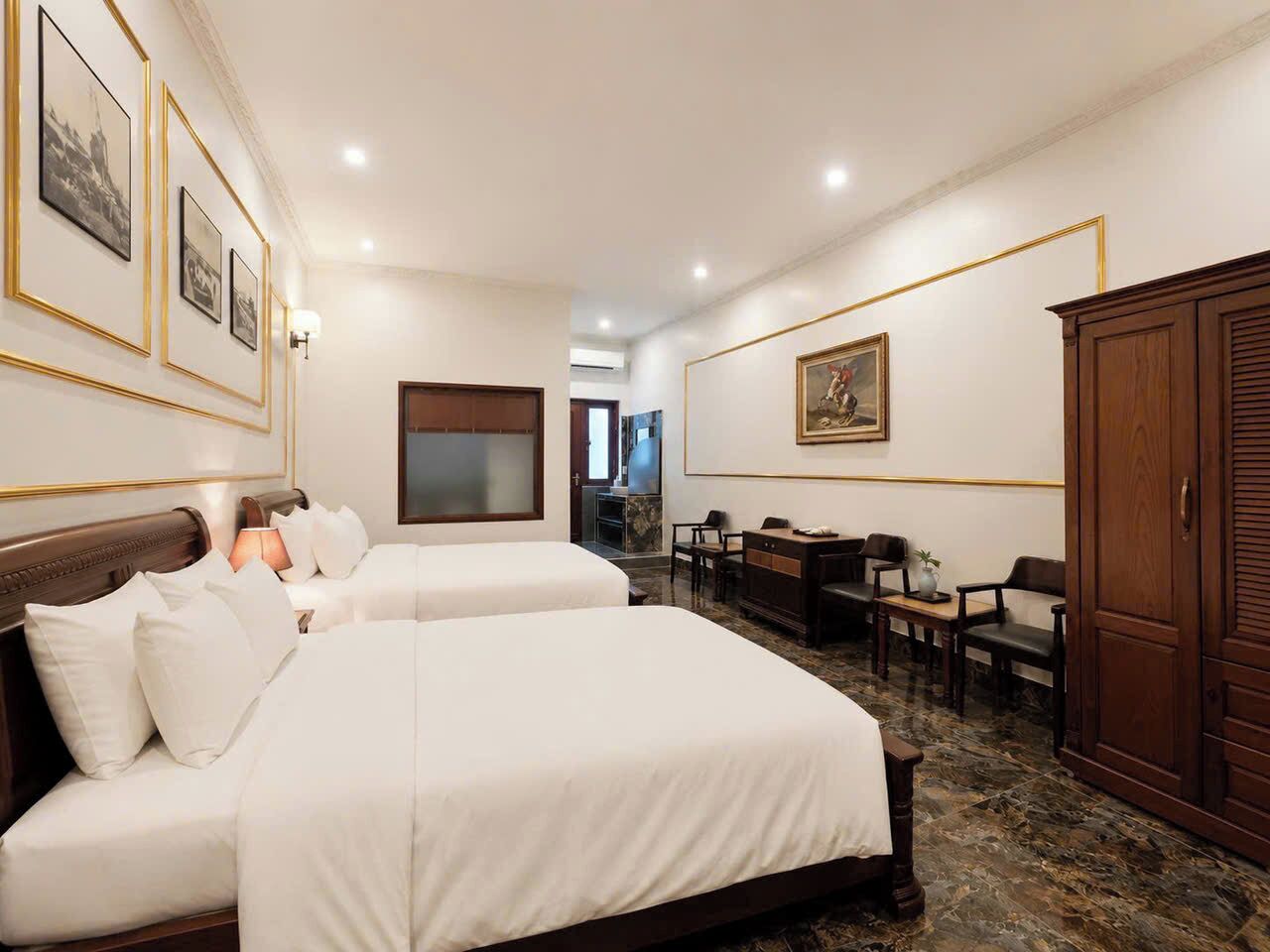 Une chambre d’hôtel avec deux lits, un bureau, des chaises, une armoire et des tableaux encadrés aux murs.