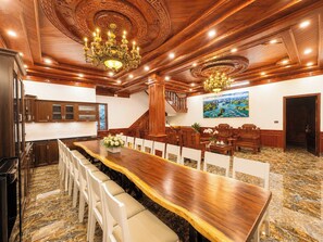 A spacious room with a long wooden table, white chairs, a chandelier, and a painting on the wall. - Rose Wood Cat Ba Villa (Hai Phong)