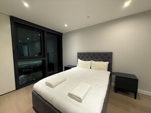 A modern bedroom with a large bed, a nightstand, a window with a city view, and a dark headboard. - Stay in Elephant and Castle (London)